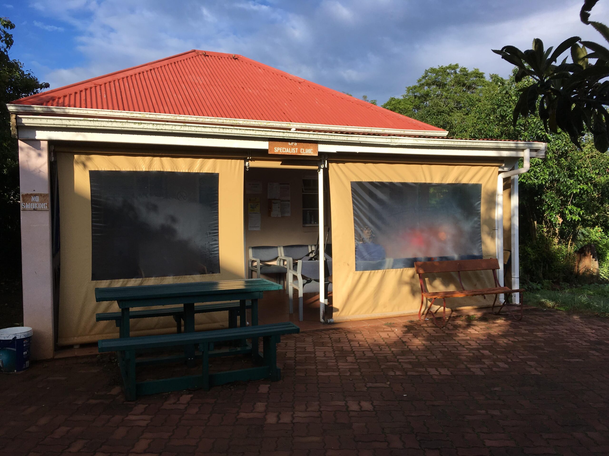 An eye-level photograph of the "Specialist Clinic" building, a small structure with a prominent red corrugated metal roof and off-white walls. The front of the clinic is largely covered by two large, tan outdoor roller blinds with rectangular clear plastic windows, which frame a central open doorway. Inside the shaded entryway, a small waiting area with several grey chairs and a bulletin board is visible. To the left of the entrance, a green wooden picnic table sits on a brick-paved patio, while a wooden bench is positioned to the right. A small "No Smoking" sign is mounted on the far-left pillar, and lush green trees and a cloudy, blue-tinted sky provide the backdrop.