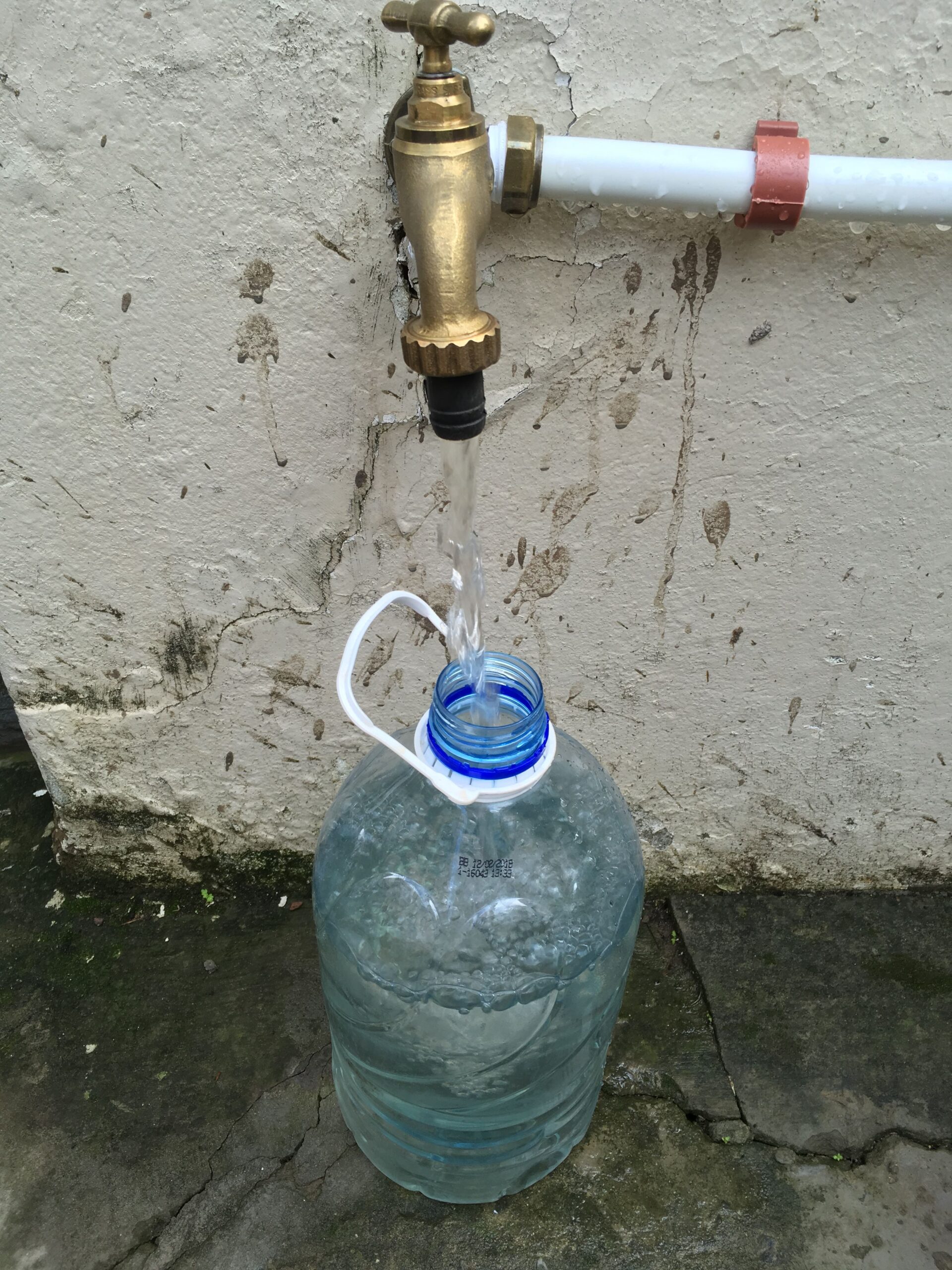 A vertical, eye-level photograph shows a steady stream of clear water flowing from a brass outdoor tap into the narrow opening of a large, clear plastic water jug. The jug, which is about half-full, sits on a cracked, dark grey concrete surface. The brass tap is connected to a white PVC pipe secured against a weathered, light-colored textured wall with a reddish-brown clip. The wall behind the tap shows signs of wear, with visible cracks and dark splatters.